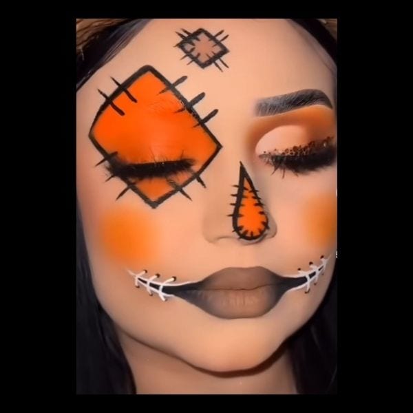 Scarecrow makeup with stitched orange patches over one eye and the forehead, an orange triangle nose and stitches across the mouth